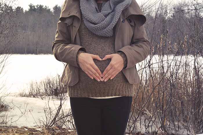 Mujer embarazada vestida con ropa de abrigo tocándose la barriga con las manos en forma de corazón.